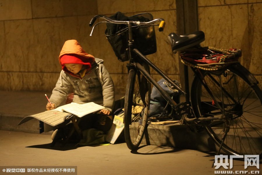 鄭州中原路京廣鐵路大橋下涵洞的馬路牙子邊，一位戴著紅色辣椒帽的小男孩趴在一個小馬扎上，上面鋪著一張車墊，在微弱的燈光下寫作業(yè)