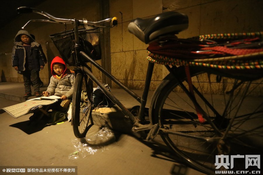 2015年11月8日晚，鄭州，7歲男孩涵洞橋下陪父母練攤，路邊燈下坐小馬扎上寫作業(yè)。