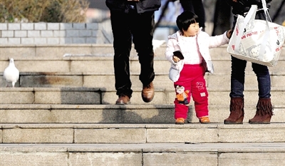 昨天中午，一女孩拉著家長的手在紫荊山公園曬太陽，氣溫升高，孩子的棉衣解開了扣子。